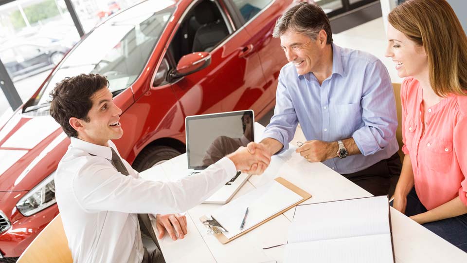Ein Autoverkäufer schüttelt den Kunden die Hand in einem Autohaus in Aschaffenburg, im Hintergrund ist ein rotes Auto zu sehen und auf dem Tisch liegen Unterlagen.